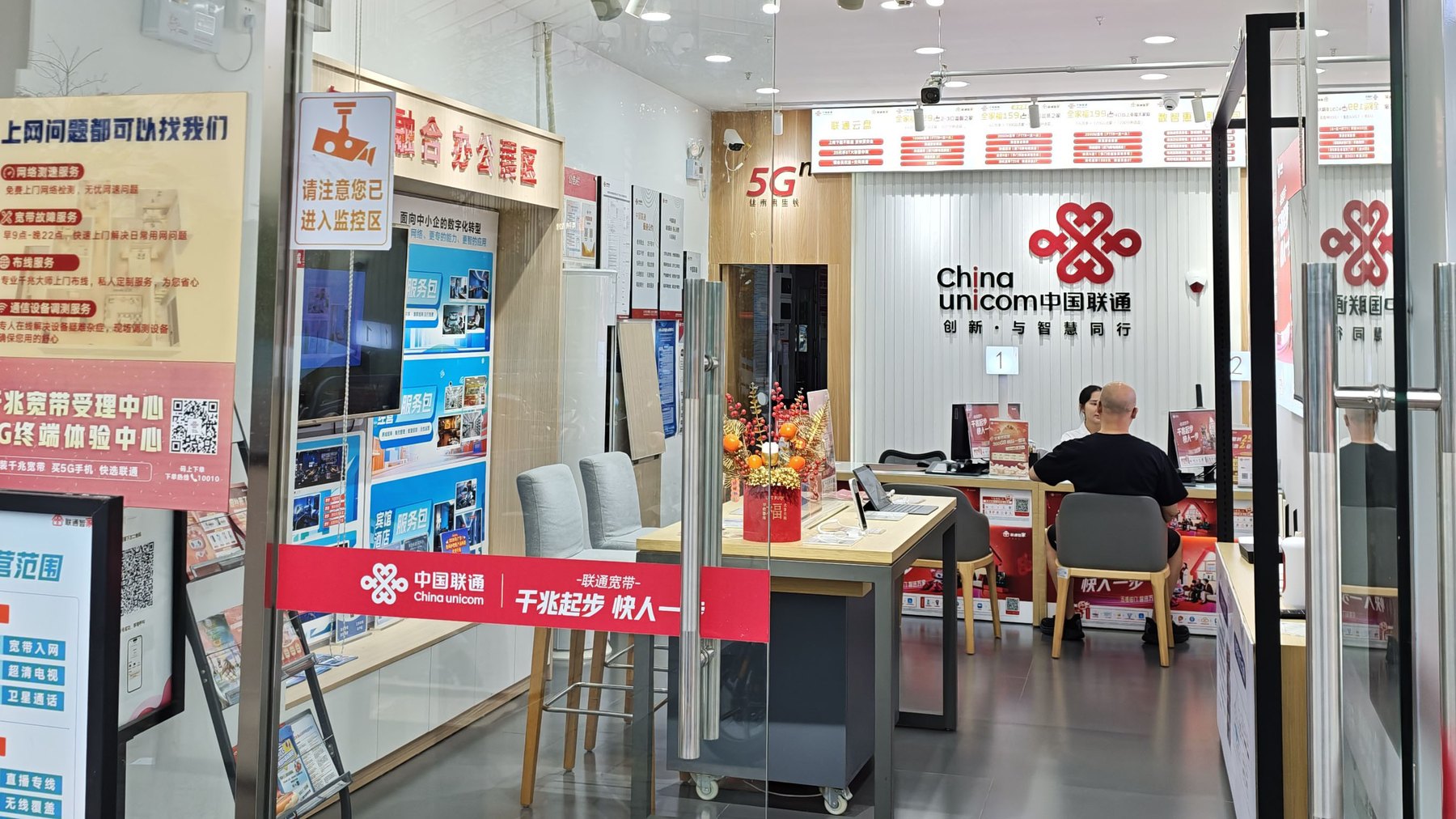 一家中国联通营业厅的店内实拍:可见“千兆宽带受理中心”“千兆起步 快人一步”等宣传物料,柜台前有顾客正在办理业务